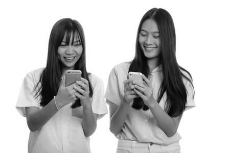 Two young happy Asian teenage girls smiling and using mobile phones togetherの写真素材