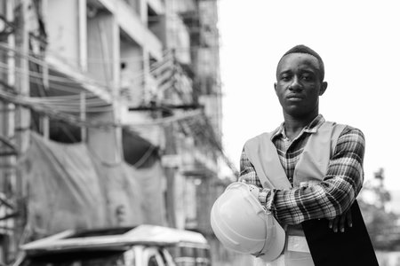 Young black African man construction worker holding clipboard and hard hat with arms crossed at...
