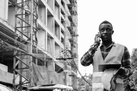 Young African man construction worker thinking while talking on the phone at building siteの写真素材
