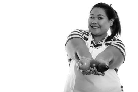 Studio shot of young happy fat Asian woman smiling while giving fresh fruits and wearing apronの写真素材