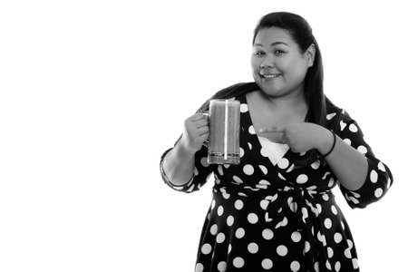 Studio shot of young happy fat Asian woman smiling while holding and pointing at glass of vegetable smoothieの写真素材