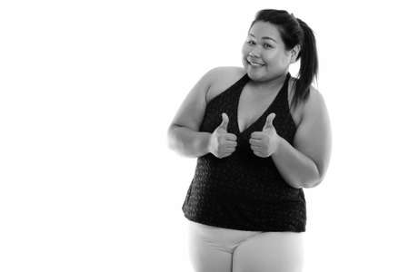 Studio shot of young happy fat Asian woman smiling and giving thumbs up ready for gymの写真素材