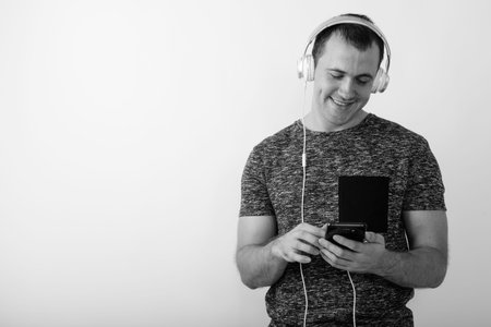 Studio shot of young happy muscular man smiling and listening to music while using mobile phone...