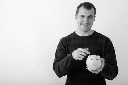 Studio shot of young happy muscular man smiling while holding and pointing at piggy bank against white backgroundの写真素材