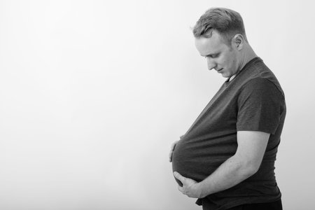 Profile view of young handsome man holding and looking at watermelon as pregnant stomach against...