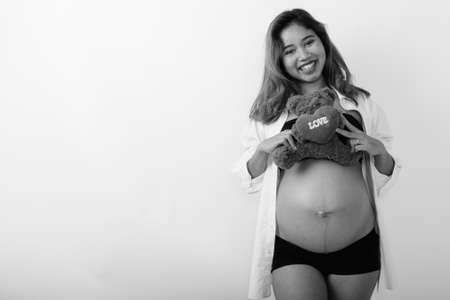 Studio shot of young happy Asian pregnant woman smiling while holding teddy bear with heart and love signの写真素材