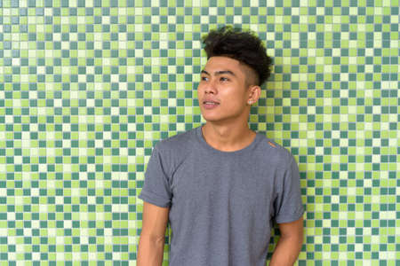 Happy young Asian man with curly hair thinking against green wall outdoorsの写真素材