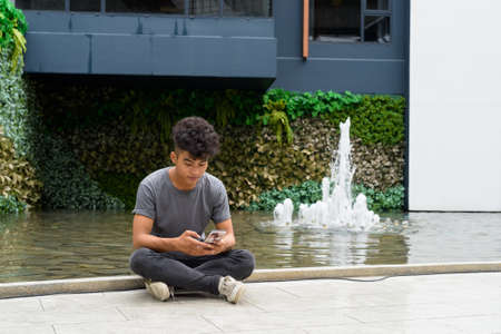 Young Asian man sitting while using phone outside modern buildingの写真素材