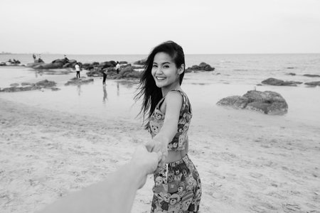 Young happy beautiful Asian woman smiling while holding hands and leading man at the public beach of...