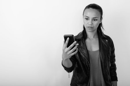 Studio shot of young Asian woman using mobile phone while wearing leather jacket against white...