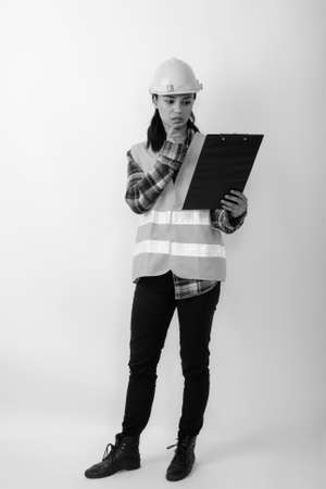 Full body shot of young Asian woman construction worker thinking while reading on clipboardの写真素材