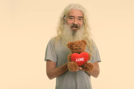 Studio shot of happy senior bearded man smiling while holding teddy bear with heart and love sign ready for Valentines dayの写真素材
