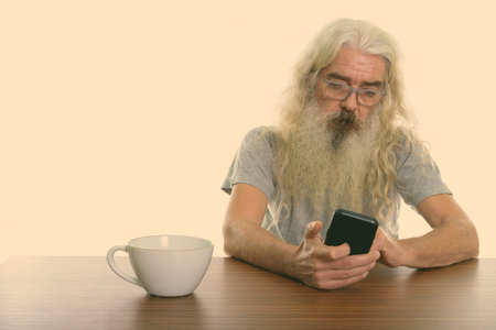 Studio shot of senior bearded man using mobile phone with coffee cup on wooden table