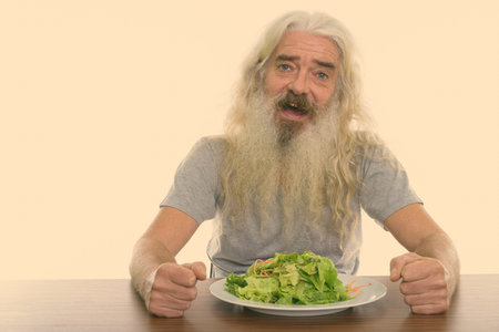Studio shot of happy senior bearded man smiling with plate of salad on wooden table