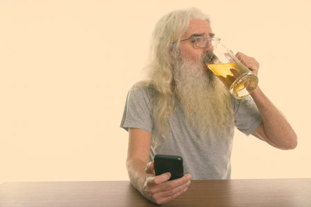 Studio shot of senior bearded man holding mobile phone and drinking glass of beer on wooden table