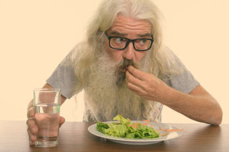 Close up of senior bearded man thinking and wearing eyeglasses while eating plate of salad with glass of water on wooden tableの写真素材