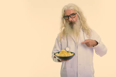 Studio shot of senior bearded man doctor holding and pointing at bowl of potato chipsの写真素材