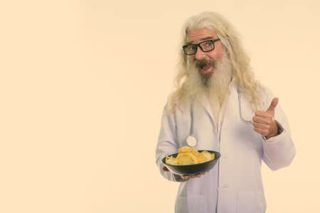 Studio shot of happy senior bearded man doctor smiling while giving thumb up and holding bowl of potato chipsの写真素材
