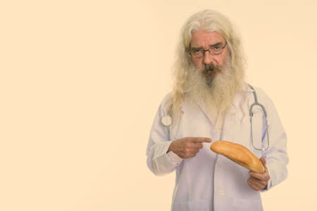 Studio shot of senior bearded man doctor wearing eyeglasses while holding and pointing at breadの写真素材