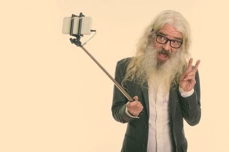 Studio shot of happy senior bearded businessman taking selfie with phone on selfie stick