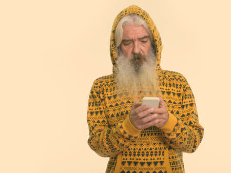 Studio shot of senior bearded man wearing hoodie while using mobile phoneの写真素材
