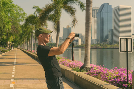 Profile view of senior tourist man wearing cap while taking pictures at peaceful park in Bangkok...