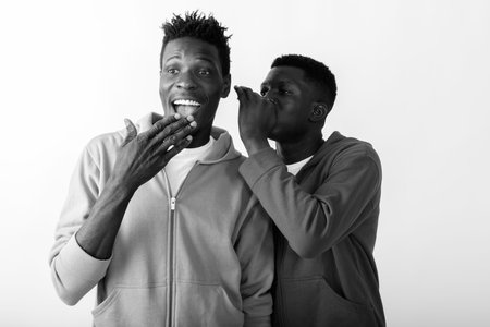 Young African man whispering to happy African man smiling while thinking and looking shocked