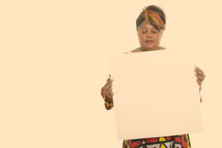 Studio shot of fat black African woman holding and looking at empty white board with copy spaceの写真素材