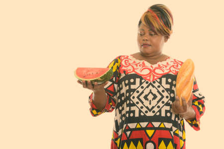 Studio shot of fat black African woman holding bread and looking at slice of watermelonの写真素材