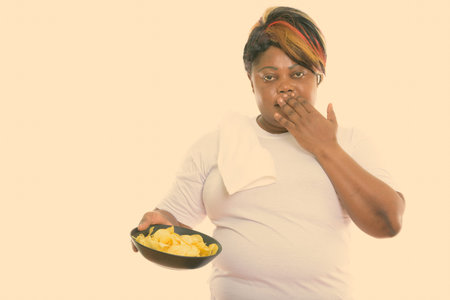 Studio shot of fat black African woman looking shocked while holding bowl of potato chips