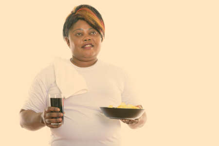Studio shot of happy fat black African woman smiling and thinking while holding glass of soda drink and bowl of potato chipsの写真素材