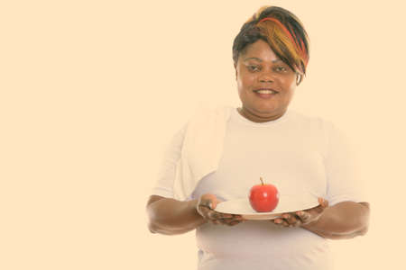 Studio shot of happy fat black African woman smiling while holding red apple served on white plate ready for gymの写真素材