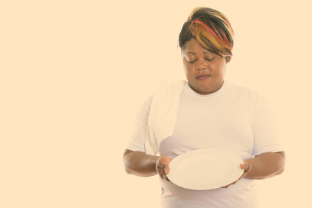 Studio shot of fat black African woman holding and looking at empty white plate with towel over the...