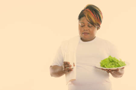 Studio shot of fat black African woman holding lettuce served on white plate and looking at glass of milkの写真素材