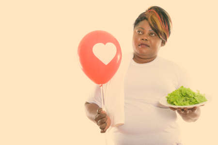 Studio shot of fat black African woman thinking while holding lettuce served on white plate and red balloon with heart signの写真素材
