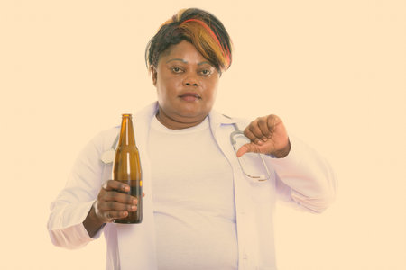 Studio shot of fat black African woman doctor holding bottle of beer while giving thumb down