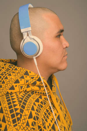 Young bald Asian man listening to music against gray backgroundの写真素材