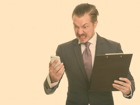 Young handsome Caucasian businessman looking angry while using mobile phone and holding clipboard