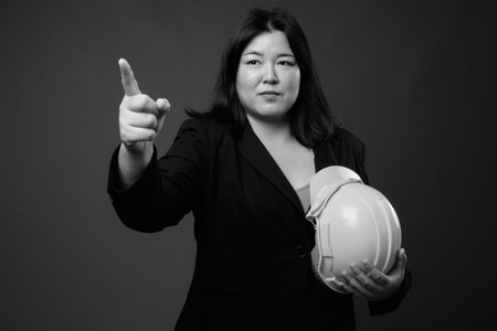 Beautiful overweight Asian businesswoman holding hardhat against gray backgroundの写真素材