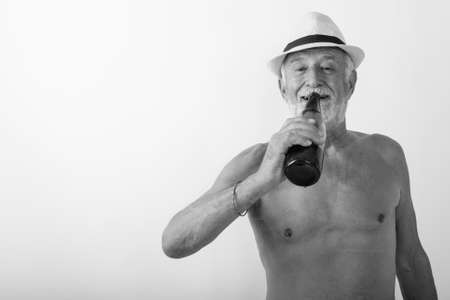 Studio shot of happy senior bearded man smiling while drinking bottle of beer and wearing hat shirtless against white backgroundの写真素材