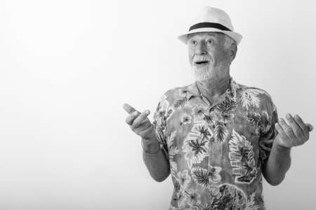 Studio shot of happy senior bearded tourist man smiling while looking surprised with both arms raised and wearing hat against white backgroundの写真素材