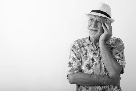 Studio shot of senior bearded tourist man thinking while looking stressed and wearing hat against...