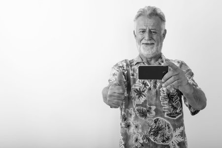 Studio shot of happy senior bearded tourist man smiling while giving thumb up and taking picture...