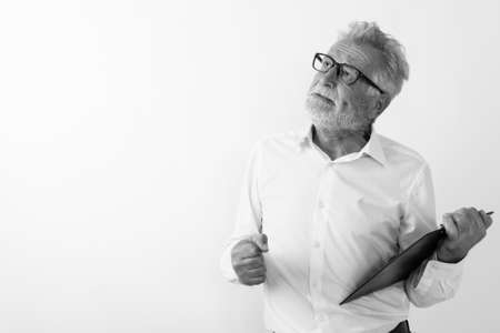 Studio shot of angry senior bearded man thinking while holding clipboard with eyeglasses against white backgroundの写真素材