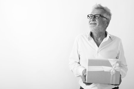 Studio shot of happy senior bearded man smiling and thinking while holding gift box with eyeglasses...