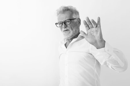Studio shot of senior bearded man looking disgusted while showing stop hand sign and wearing...