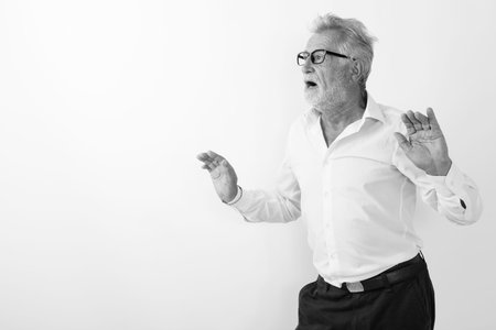 Studio shot of handsome senior bearded man looking surprised with both arms raised while wearing eyeglasses against white backgroundの写真素材