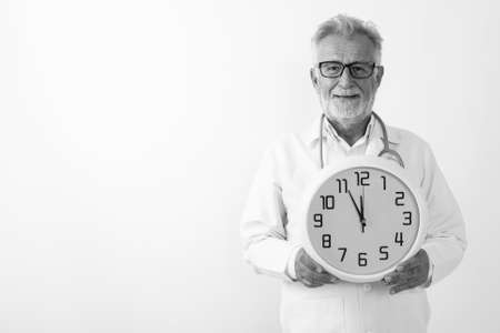 Studio shot of happy senior bearded man doctor smiling while holding wall clock against white backgroundの写真素材