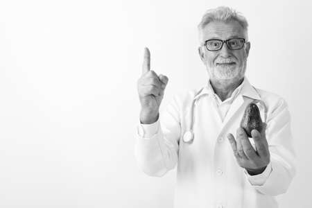 Studio shot of handsome senior bearded man doctor holding avocado while pointing finger up against white backgroundの写真素材