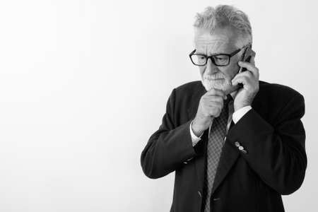Studio shot of handsome senior bearded businessman thinking while talking on mobile phone against white backgroundの写真素材